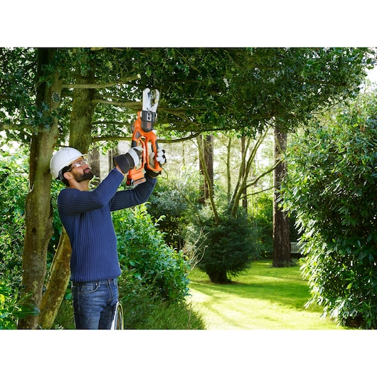 A person wearing protective gear is using an orange and black BLACK+DECKER cordless garden pruner to trim branches on a tree in a garden.