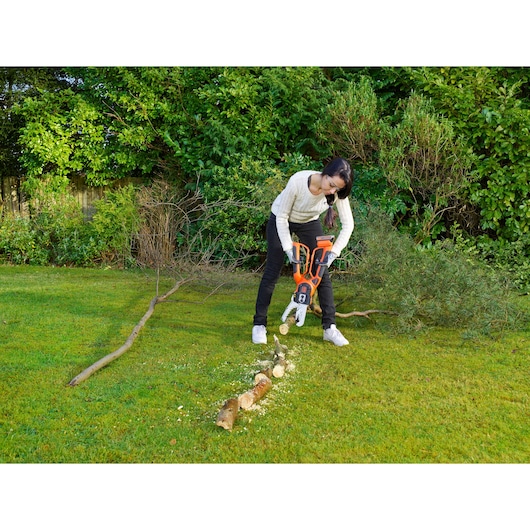 A person using a BLACK+DECKER GKC1000L cordless chainsaw to cut branches on a lawn, surrounded by green bushes and trees in a garden.