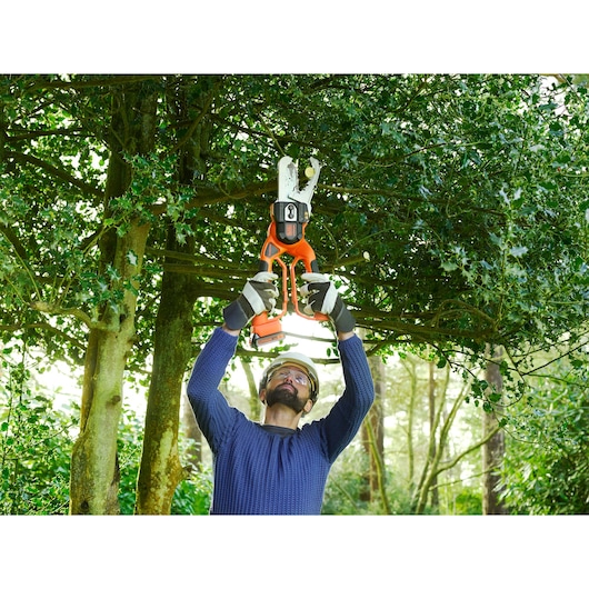 A person wearing protective gloves and a helmet is using an orange BLACK+DECKER cordless power pruner to cut branches from a tree in a garden or forest setting.