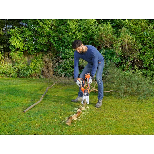 A person using a BLACK+DECKER GKC1000L cordless chainsaw to cut a tree branch on a grassy lawn, with dense greenery in the background. The user is wearing gloves and casual clothing.