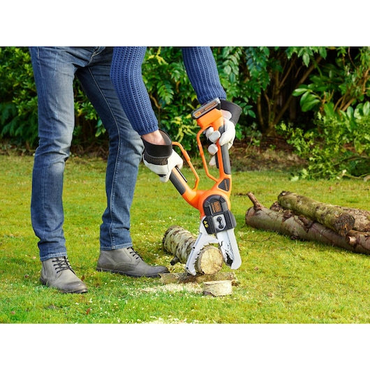 A person using a BLACK+DECKER cordless chainsaw (model GKC1000L) to cut a small log outdoors on grass, wearing gloves and casual clothing. The log is placed on the ground with other logs in the background.