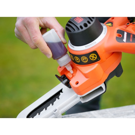 Close-up of a person adding oil to a BLACK+DECKER GKC1000L battery-powered chainsaw, with orange housing and safety symbols visible.