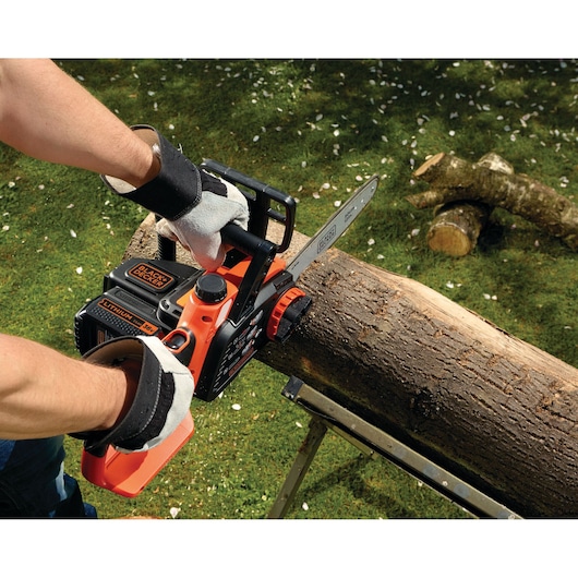 A person wearing protective gloves is using a BLACK+DECKER cordless chainsaw to cut a log outdoors, with another log visible on the grass nearby.