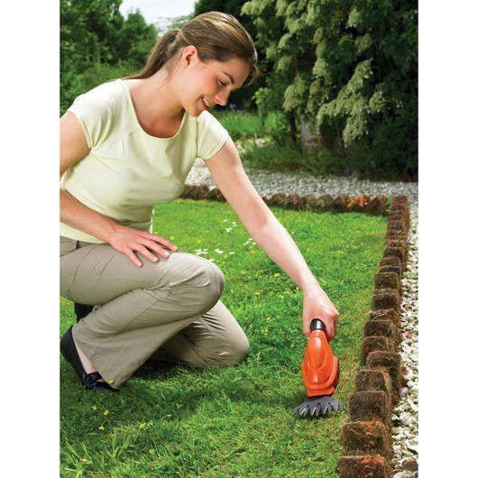 A person kneeling in a garden uses a BLACK+DECKER GSL200 cordless grass shear to trim the grass along the edge of a lawn bordered with bricks.