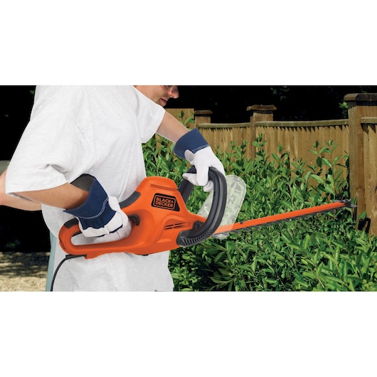 A person wearing gloves is using an orange BLACK+DECKER electric hedge trimmer to cut hedges near a wooden fence.