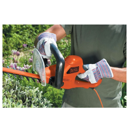 A person wearing protective gloves is using an orange BLACK+DECKER electric hedge trimmer in a garden, trimming green bushes.