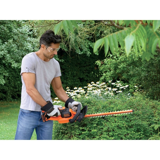 A person using a BLACK+DECKER cordless hedge trimmer to trim bushes in a garden. The person is wearing gloves and a short-sleeved shirt.