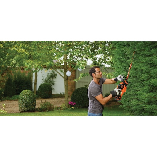 A person using a BLACK+DECKER cordless hedge trimmer (model GTC18504PC) to trim a tall green hedge in a garden. The person is wearing gloves and a grey shirt, and the garden features trimmed bushes and trees in the background.