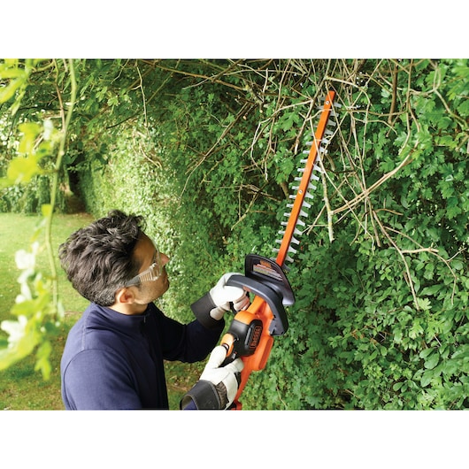 A person wearing gloves is using a BLACK+DECKER GTC18504PC cordless hedge trimmer to trim dense green hedges outdoors. The individual's face is blurred for privacy.