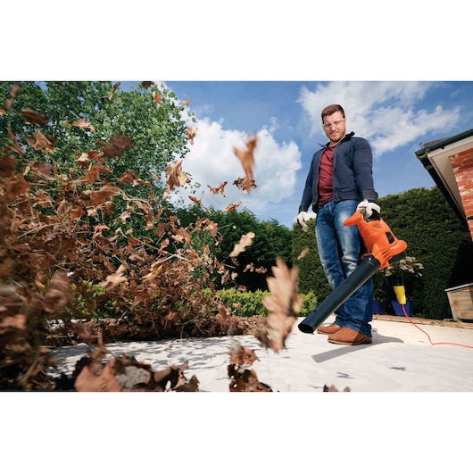 A person using an orange BLACK+DECKER leaf blower to clear dried leaves from a garden area. The person is wearing gloves, jeans, and a jacket, and the leaves are being blown away energetically.
