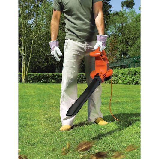 A person wearing gloves uses an orange BLACK+DECKER electric leaf blower in a garden to clear leaves from the grass.
