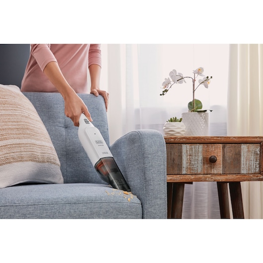 BLACK+DECKER HLVC315B11 handheld vacuum cleaning crumbs from blue sofa near wooden side table.