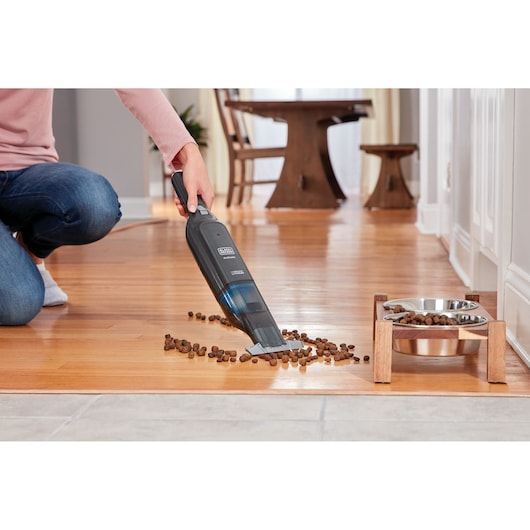 BLACKDECKER HLVC320B11 handheld vacuum cleaning pet food on wood floor near dog bowls.
