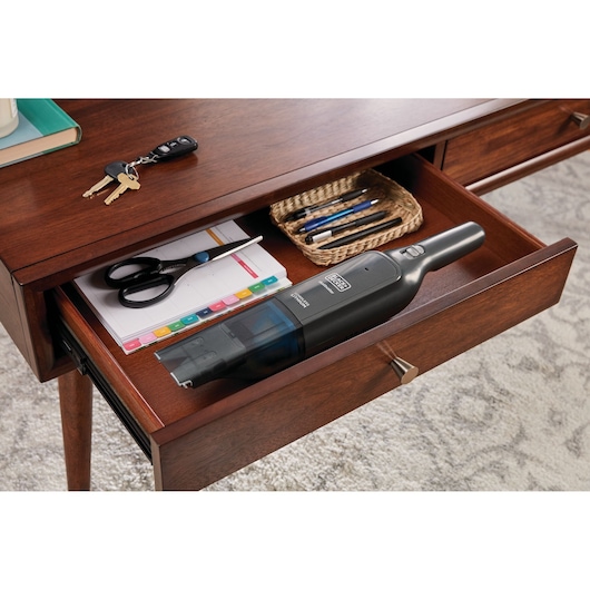 Photo of BLACK+DECKER HLVC320B11 handheld vacuum cleaner in an open wooden desk drawer with office supplies.