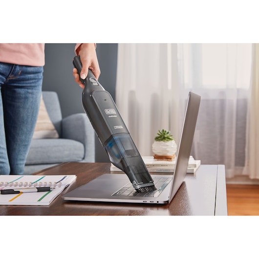 Photo of BLACK+DECKER HLVC320J11 handheld vacuum cleaning a laptop keyboard on a wooden desk in a living room.