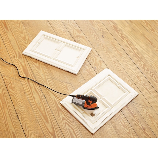 BLACKDECKER KA2000 detail sander on white cabinet door panel, wood floor background, angled top view.