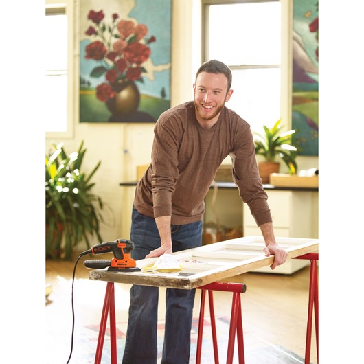 A person with a blurred face is working indoors on a wooden surface, using a BLACK+DECKER KA2500K power tool placed on a table supported by red sawhorses. Art and plants decorate the background.