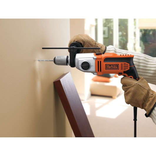 A person wearing gloves uses a BLACK+DECKER electric drill to make a hole in a beige wall indoors. The drill is orange and black, and the environment appears to be a living room.