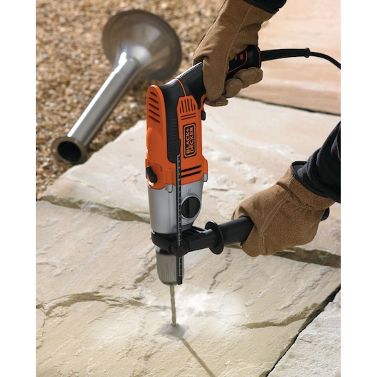 A person wearing protective gloves is using a BLACK+DECKER electric drill to make a hole in a stone or concrete surface outdoors. A metal nozzle or tube is visible in the background.