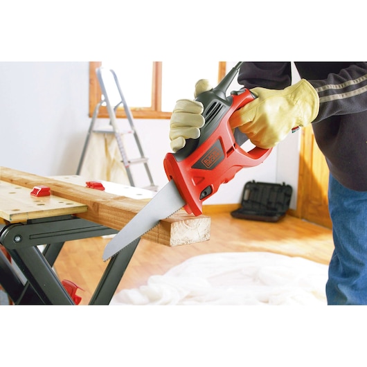 A person wearing yellow work gloves is using a BLACK+DECKER reciprocating saw to cut a piece of wood placed on a workbench. A ladder and a toolbox are visible in the background.