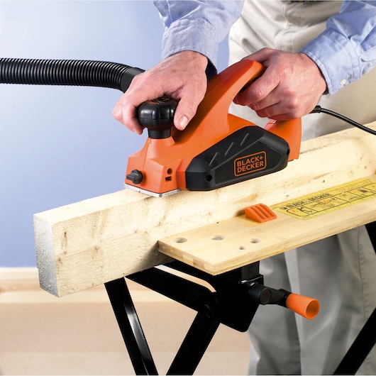 Photo of BLACK+DECKER KW712 electric hand planer in use on a wooden beam, showing orange body and black accents.
