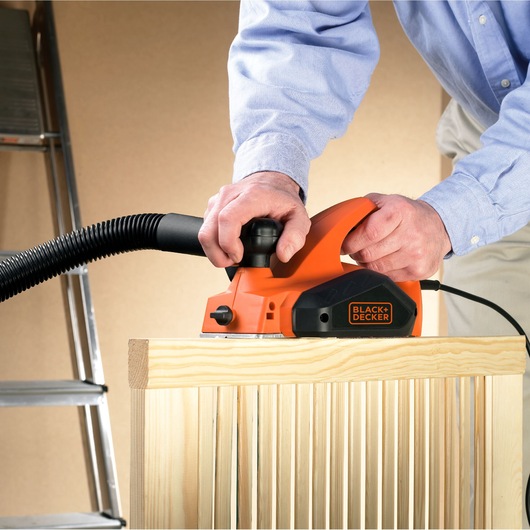 Photo of BLACK+DECKER KW712 electric planer in use on light wood, showing dust extraction hose attachment.