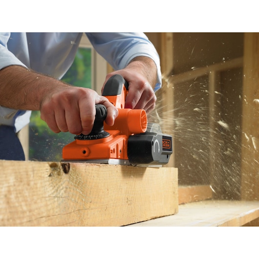 A person using an orange BLACK+DECKER electric planer to smooth a piece of wood, with wood shavings flying in a workshop setting.