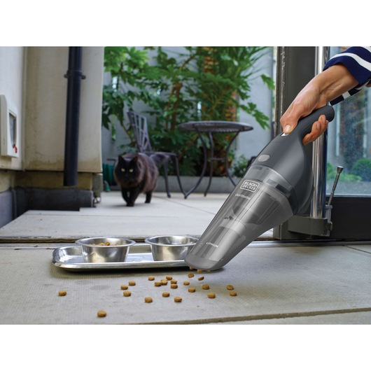 BLACK+DECKER NVB215WA handheld vacuum cleaning pet food near metal bowls on patio floor with a black cat in the background.
