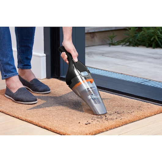 A person uses a BLACK+DECKER handheld vacuum cleaner to remove dirt from a doormat near the entrance of a house.
