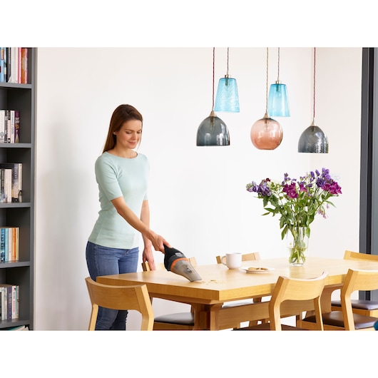 A person using a BLACK+DECKER handheld vacuum cleaner to clean crumbs from a wooden dining table. The table is surrounded by chairs and has flowers in a vase, a cup, and a plate on it. Several colorful pendant lights hang above the table, and there is a bookshelf in the background.