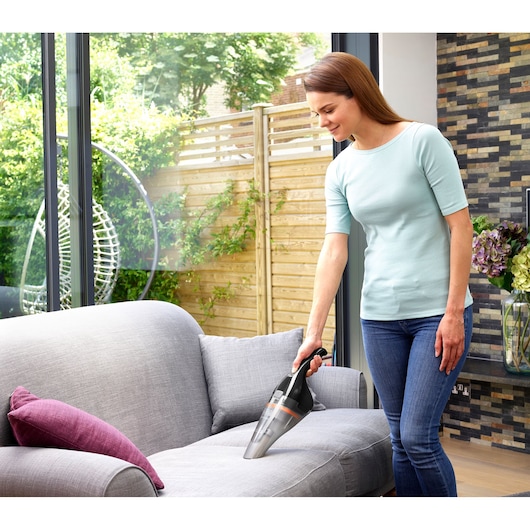 A person using a BLACK+DECKER handheld vacuum cleaner (model NVC115BJL) to clean a gray sofa in a modern living room with large windows and greenery visible outside.