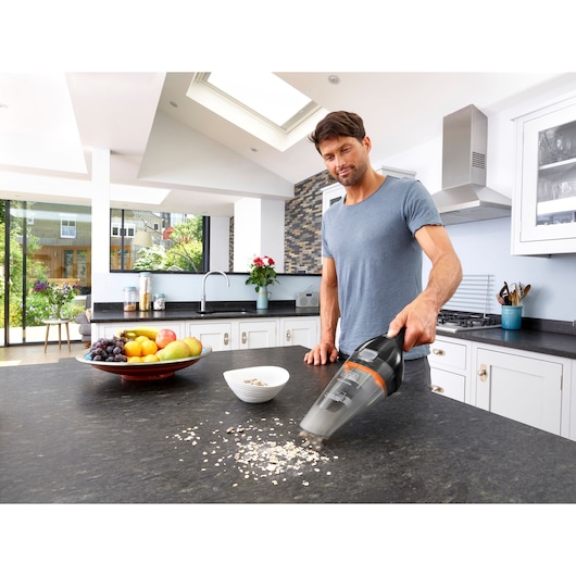 A person using a BLACK+DECKER handheld vacuum cleaner model NVC115BJL to clean spilled oats on a kitchen countertop. There is a bowl and a plate of fruit nearby. The kitchen is modern and bright.