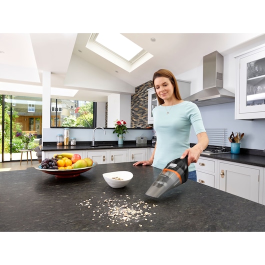 A person using a BLACK+DECKER handheld vacuum cleaner (model NVC115BJL) to clean spilled cereal on a kitchen countertop. The kitchen is modern with large windows, a fruit bowl, and a bowl of cereal on the counter.