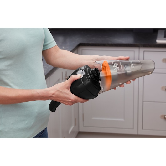 A person holding and opening a BLACK+DECKER handheld vacuum cleaner in a kitchen setting, showing the dust container and filter.