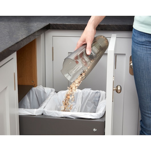 A person empties debris from a BLACK+DECKER handheld vacuum cleaner into a trash bin lined with a plastic bag, inside a kitchen cabinet.