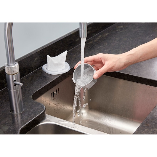 A person rinsing a plastic filter under running water from a kitchen faucet, with another filter placed on the kitchen counter beside the sink.