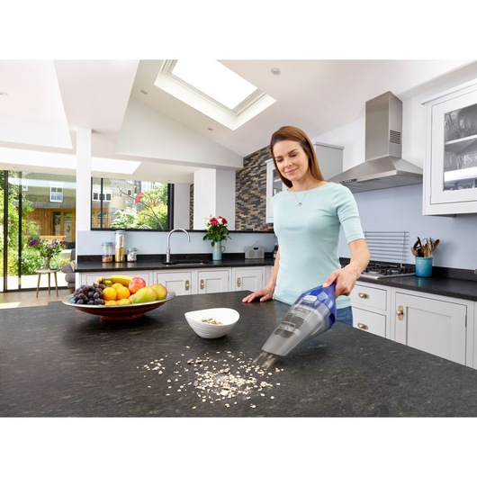 A person using a BLACK+DECKER NVC115WA handheld vacuum cleaner to clean food debris from a kitchen countertop. A bowl of fruit and a small white bowl are also visible on the counter in a modern kitchen setting.