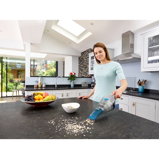 A person using a BLACK+DECKER handheld vacuum cleaner to clean up spilled oats on a kitchen countertop. There is a bowl of oats and a plate of fruit on the counter in a bright modern kitchen.