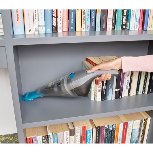 A handheld BLACK+DECKER vacuum cleaner being used to clean a gray bookshelf, with books arranged on the shelves.