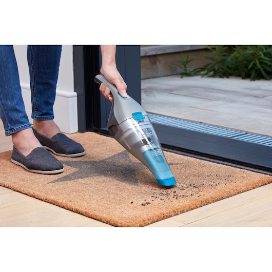 A person using a BLACK+DECKER handheld vacuum cleaner to remove dirt from a doormat near an open doorway.
