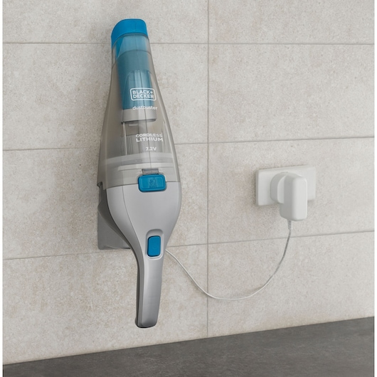 A BLACK+DECKER cordless handheld vacuum cleaner, model NVC215WA, mounted on a wall and plugged into a nearby electrical outlet for charging. The vacuum has a gray and blue design and is placed against light-colored tiled walls.