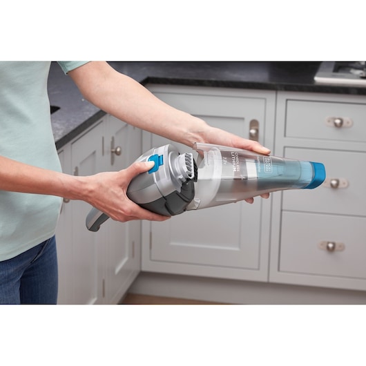 A person is holding a BLACK+DECKER NVC215WA handheld vacuum cleaner in a kitchen, demonstrating how to open and remove the dust container.