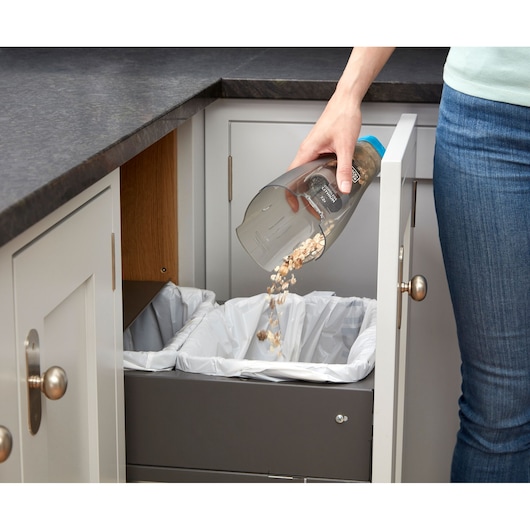 A person empties debris from a handheld vacuum cleaner into a kitchen trash bin with a pull-out drawer.