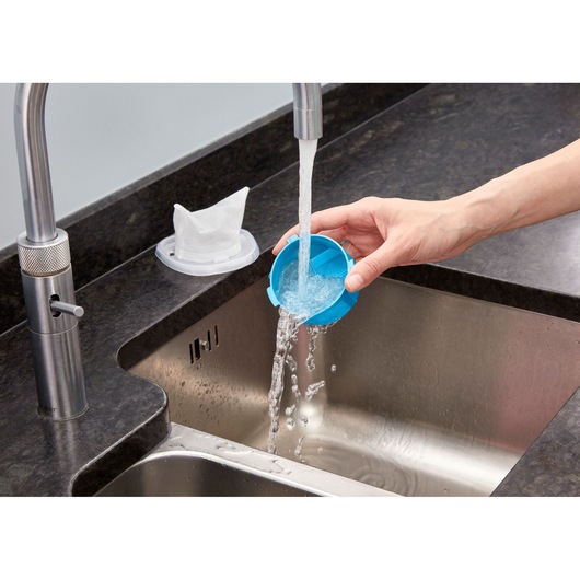 A hand rinsing a blue vacuum filter under running water from a kitchen faucet, with a white filter drying on a holder nearby on the countertop.