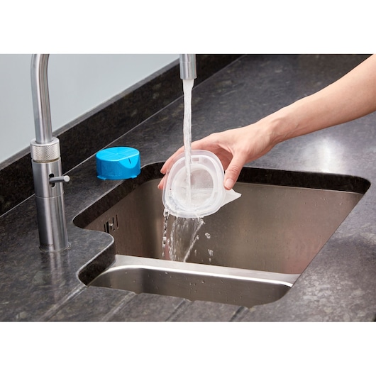 A person rinses a vacuum cleaner filter under running water at a kitchen sink, with a blue container on the counter nearby.