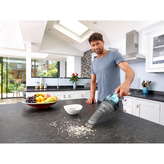 A person using a BLACK+DECKER handheld vacuum cleaner to clean spilled oats from a kitchen countertop. The kitchen features a bowl of fruit and modern decor.