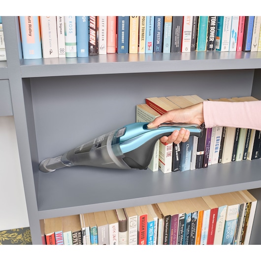 A person uses a BLACK+DECKER handheld vacuum to clean a gray bookshelf, removing dust from the area beside neatly arranged books.