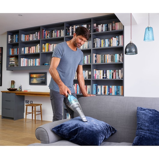 A person using a BLACK+DECKER handheld vacuum cleaner (model NVC220WBC) to clean a blue cushion on a grey sofa in a modern living room with bookshelves in the background.