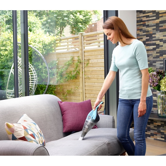 A person using a BLACK+DECKER handheld vacuum cleaner to clean a grey sofa in a modern living room. There are colorful cushions on the sofa and large windows in the background.