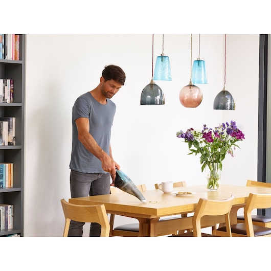 A person using a BLACK+DECKER handheld vacuum to clean crumbs from a wooden dining table in a modern room with colorful pendant lights and a vase with purple flowers. The person's face is blurred for privacy.